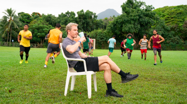 man sits in white chair on a field with soccer players in NEXT GOAL WINS