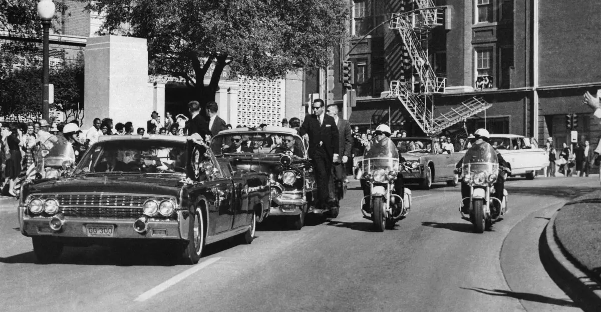 Seen through the foreground convertible's windshield, President John F. Kennedy's hand reaches toward his head within seconds of being fatally shot as First Lady Jacqueline Kennedy holds his forearm as the motorcade proceeds along Elm Street past the Texas School Book Depository in Dallas, on Nov. 22, 1963. (James W. "Ike" Altgens/AP Photo)