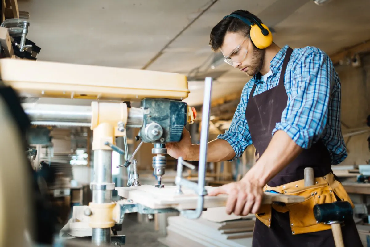 Carpenter uses a drill press on wooden board while standing in workshop. (Shutterstock)