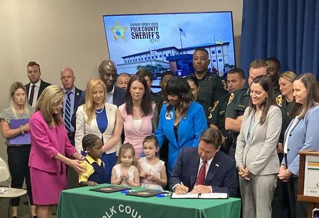 Florida Gov. Ron DeSantis signs a bill making it a felony to be in Florida after being deported with (L-to-R) Nikki Jones (in pink) and her daughters, Rep. Kiyan Michael (in blue), Lt. Gov. Jeanette Nuñez (in gray), Attorney General Ashley Moody (light blue) before a background of deputies at the Polk County Sheriff’s Office in Winter Haven, Fla., on March 15, 2024. (John Haughey/The Epoch Times)