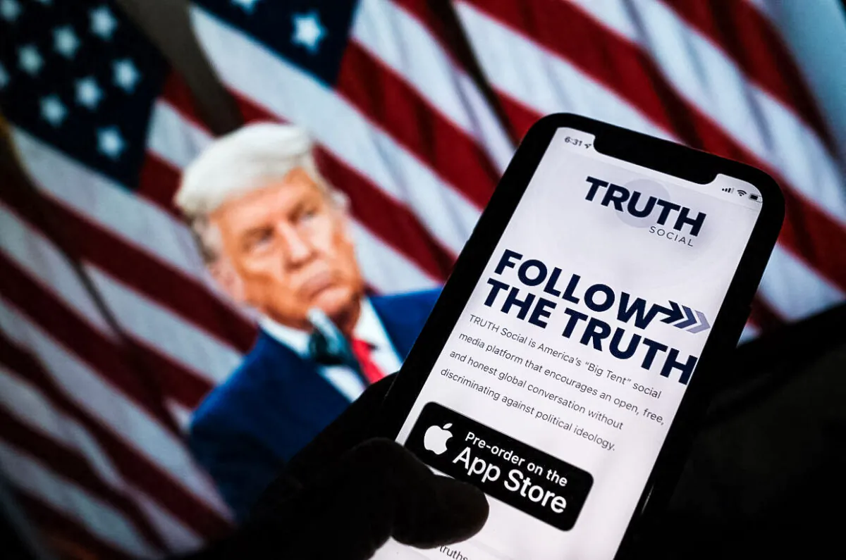 This illustration photo shows a person checking the app store on a smartphone for Truth Social with a photo of former President Donald Trump on a computer screen in the background, in Los Angeles, October 20, 2021. (Chris Delmas/AFP via Getty Images)