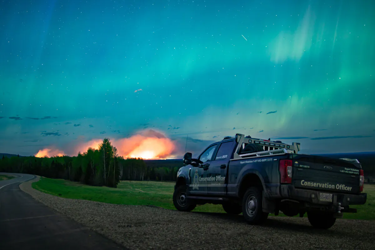 The Aurora Borealis shines overhead of a B.C. Conservation Officer Service vehicle near the junction of highways 97 and 77, as a wildfire burns in the background near Fort Nelson, B.C., in a May 11, 2024, handout photo. (The Canadian Press/HO-Ministry of Water, Land and Resource Stewardship)