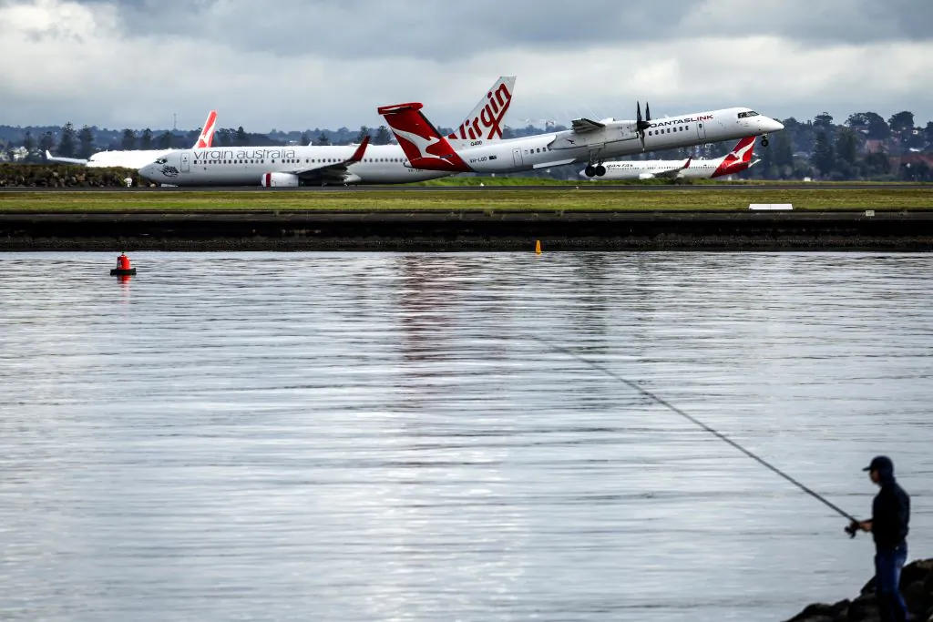 A fisherman stands on rocks as Qantas Airlines and Virgin Australia Airways planes travel down the runway at Sydney International Airport on June 7, 2024. (Photo by DAVID GRAY / AFP) (Photo by DAVID GRAY/AFP via Getty Images)