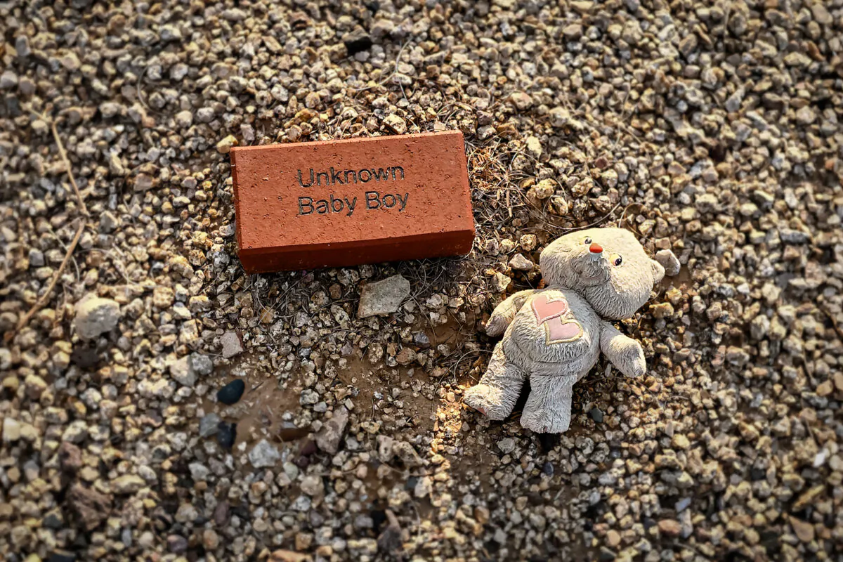 A teddy bear lies next to the brick grave marker of an unknown baby boy in White Tanks Cemetery in Goodyear, Ariz., on July 11, 2024. (Allan Stein/The Epoch Times)