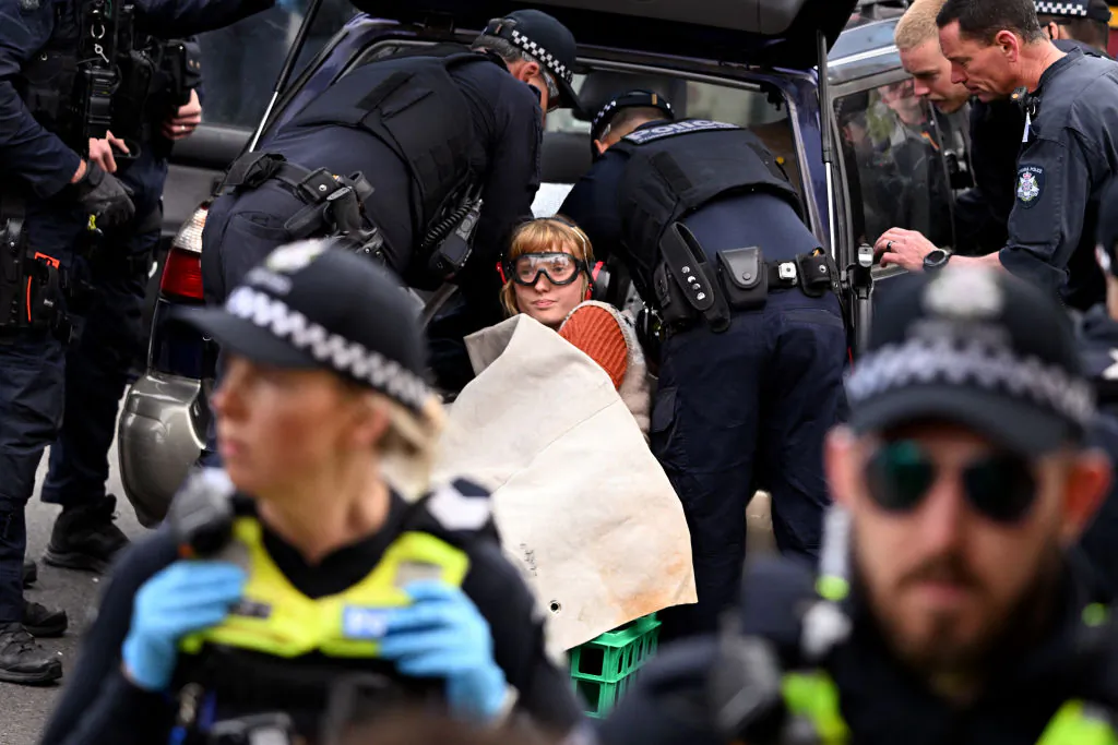 Police officers put protective gear on a protester blocking a road as they try to remove her restraints from a car she attached herself to, outside the Land Forces 2024 arms fair in Melbourne on Sept. 13, 2024. (William West/AFP via Getty Images)