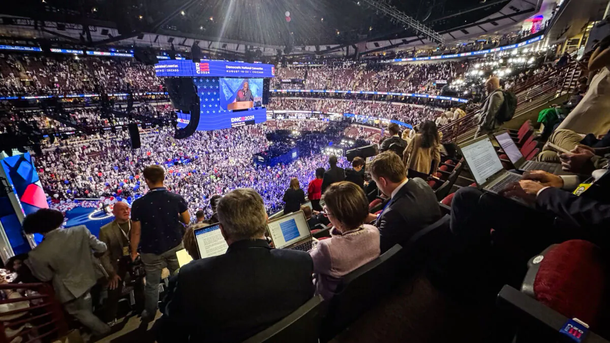 Epoch Times reporters Lawrence Wilson and Emel Akan, and national editor Ivan Pentchuokov work behind glowing screens of laptop computers in a darkened upper seating area during the Democratic National Convention in Chicago on Aug. 22, 2024. (Jan Jekielek/The Epoch Times)