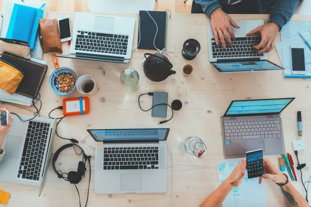 A meeting table with multiple laptops, phones and other electronic devices on top. There are also coffee cups and notebooks.  Shows people working at the table. (Marvin Meyer/UnSplash)