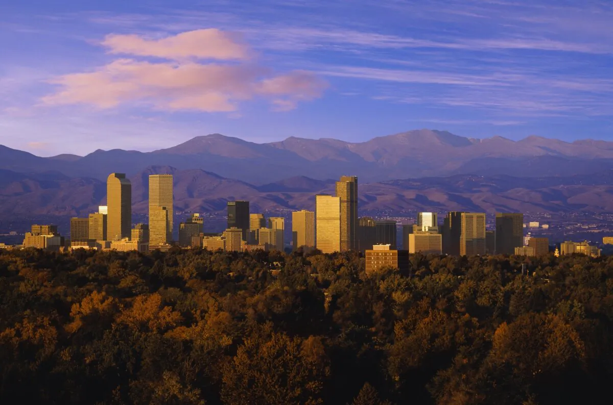 A scenic view of the Denver skyline at sunset, with the Rocky Mountains in the background and a forest in the foreground. (Bill Ross/Getty Images)