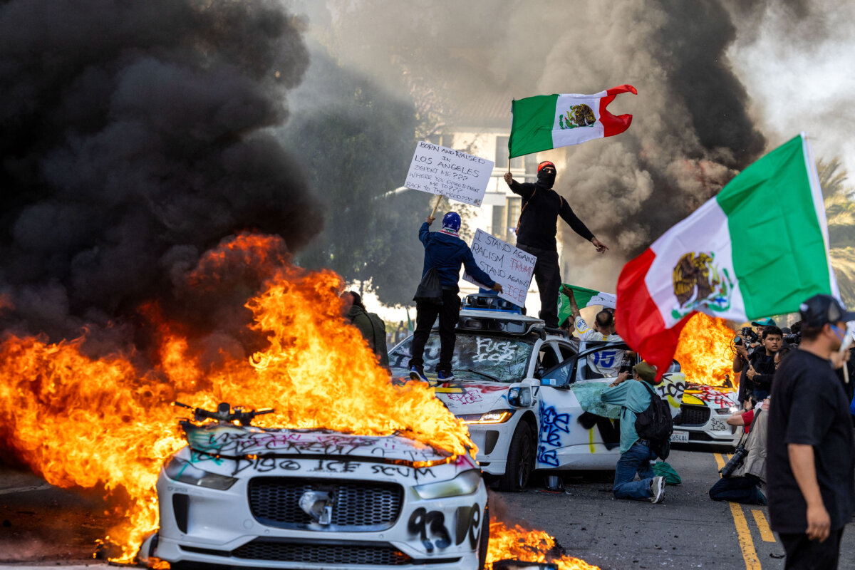 Demonstrators wave flags from atop a wrecked car, standing beside burning Waymo vehicles, as protesters clash with law enforcement in the streets near the federal building during a protest over federal immigration operations in Los Angeles on June 8, 2025. (Ringo Chiu/AFP via Getty Images)