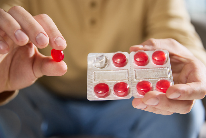 Close up photo of man in casual beige shirt holds blister pack of red lozenges, sitting in bright, cozy living room. Concept of illness, healthy insurance, pharmaceutical, medicine. Ad.