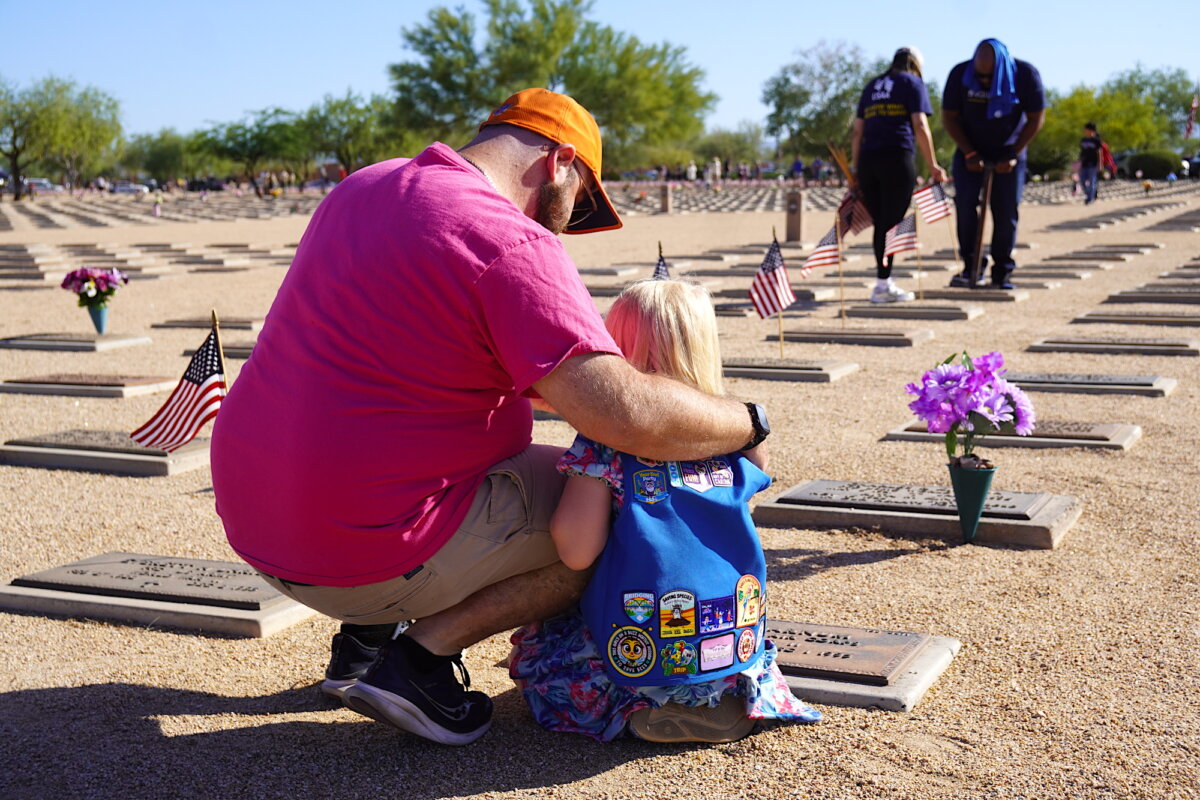 Brian Barnum (L), of Phoenix, hugs daughter Elizabeth, 5, as she visits the grave marker of her great-grandfather George Barnum, a WWII veteran, for the first time at the National Memorial Cemetery of Arizona in Phoenix on May 23, 2026. (Allan Stein/The Epoch Times)