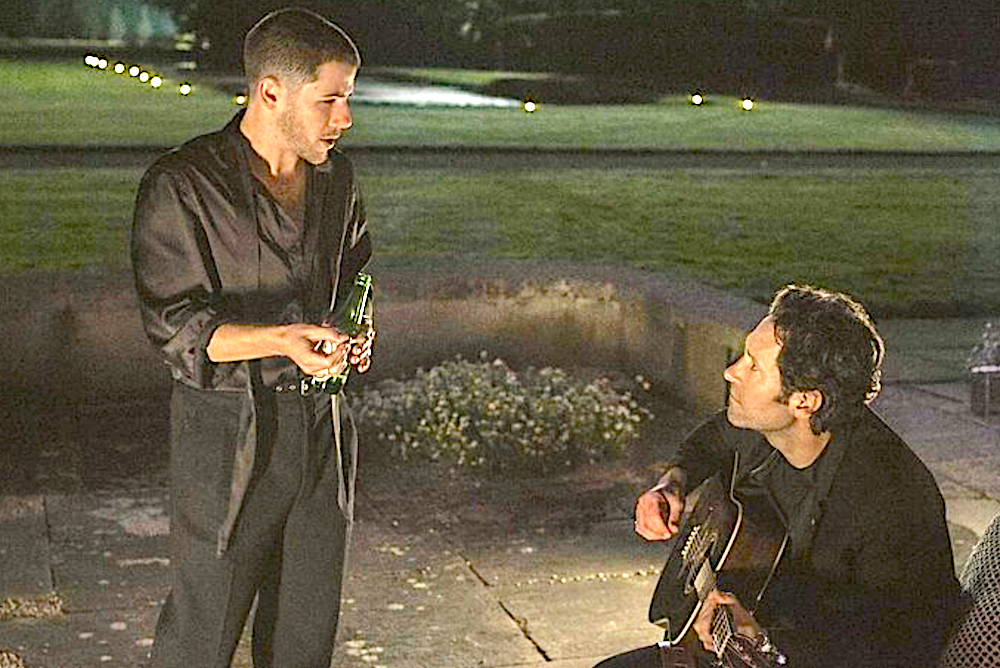 standing man talks to seated man holding a guitar in Power Ballad