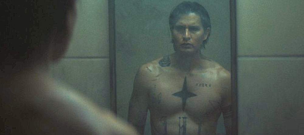 Young man with prison tattoos stands in front of mirror in Gangland