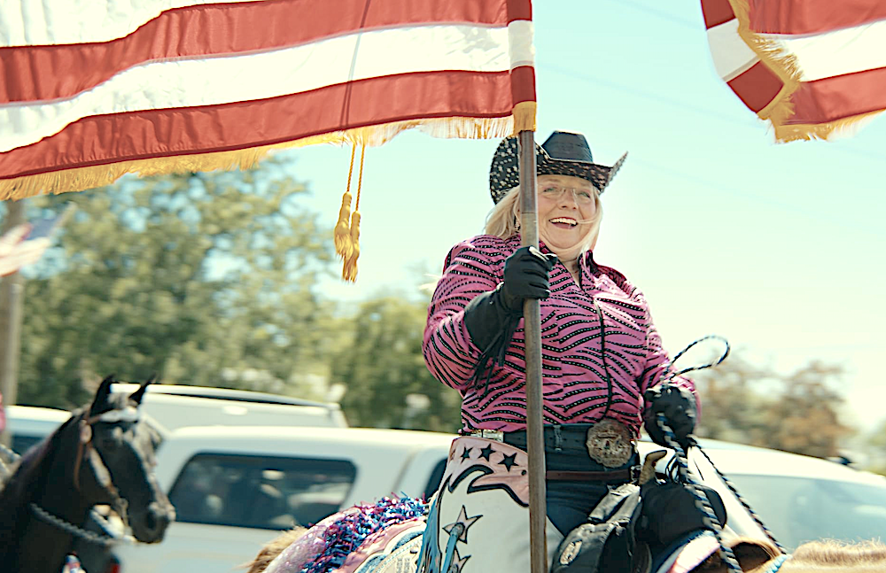 woman in pink, zebra-striped outfit with black hat, carries the flag while riding a horse, in Flag Day