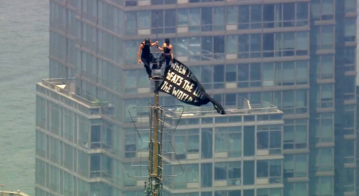 Two people stand on the tip of the antenna of the Empire State Building in New York while holding a banner on July 1, 2026. (WABC-TV via AP)
