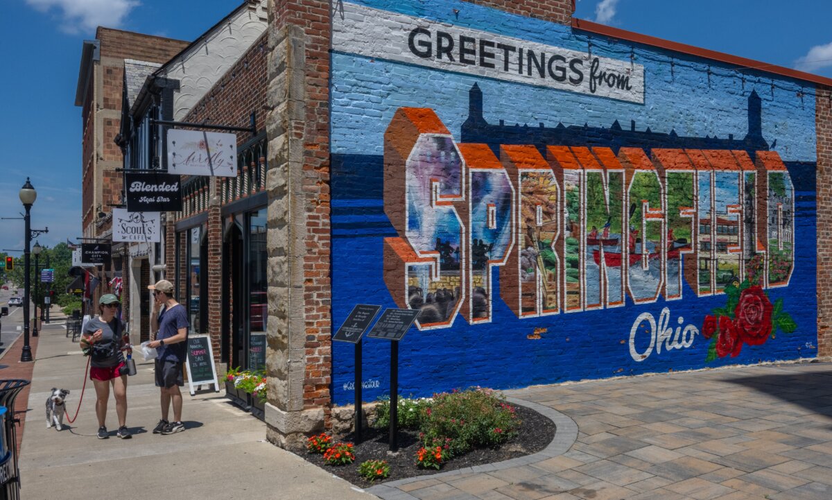 Two pedestrians and their dog walk past the Greetings from Springfield Ohio mural in Springfield, Ohio, on July 3, 2026. (Glenn Hartong for The Epoch Times)