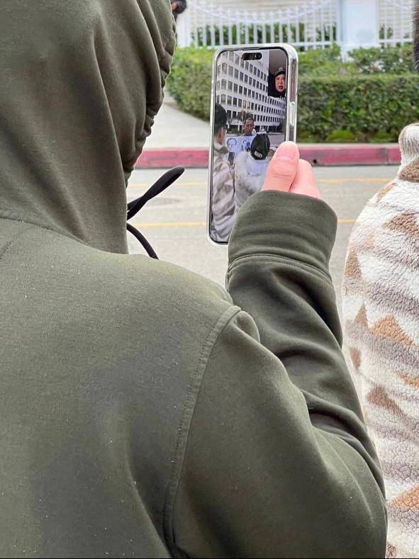 A hooded man livestreams Jie Lijian during a protest outside the Chinese Consulate in Los Angeles. The phone screen shows a Chinese man wearing a police uniform on the other end of the call. (Courtesy of Jie Lijian