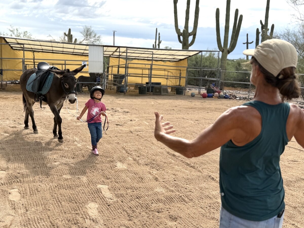 Sophie Rainer (R), equine-therapy instructor and founder of Donkeys of the Heart in Cave Creek, Ariz., guides Dylan, 4, through her riding lesson on Sept. 24, 2026. (Allan Stein/The Epoch Times)